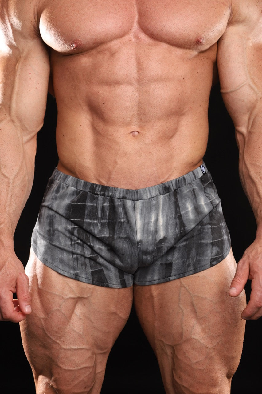 A close-up of a muscular male model wearing athletic shorts in a black acid wash color, set against a solid black background.