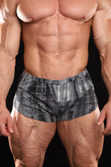 A close-up of a muscular male model wearing athletic shorts in a black acid wash color, set against a solid black background.