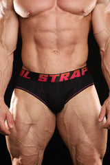 Close-up of a muscular man wearing black briefs with red text on the waistband reading 'STRAPP METAL' against a black background.