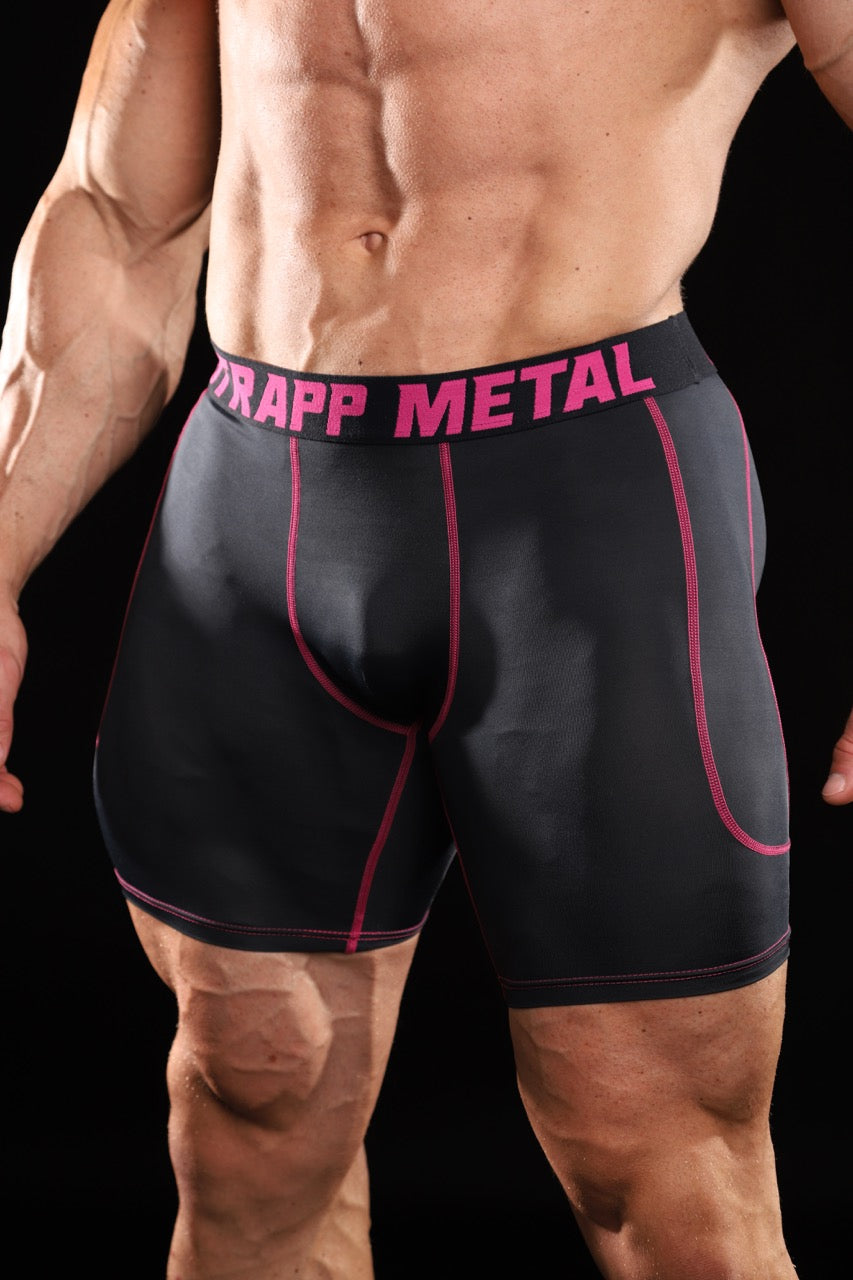 A close-up of a muscular man wearing black compression-style boxer briefs with bright pink piping and a pink and black waistband that reads 'STRAPP METAL'.